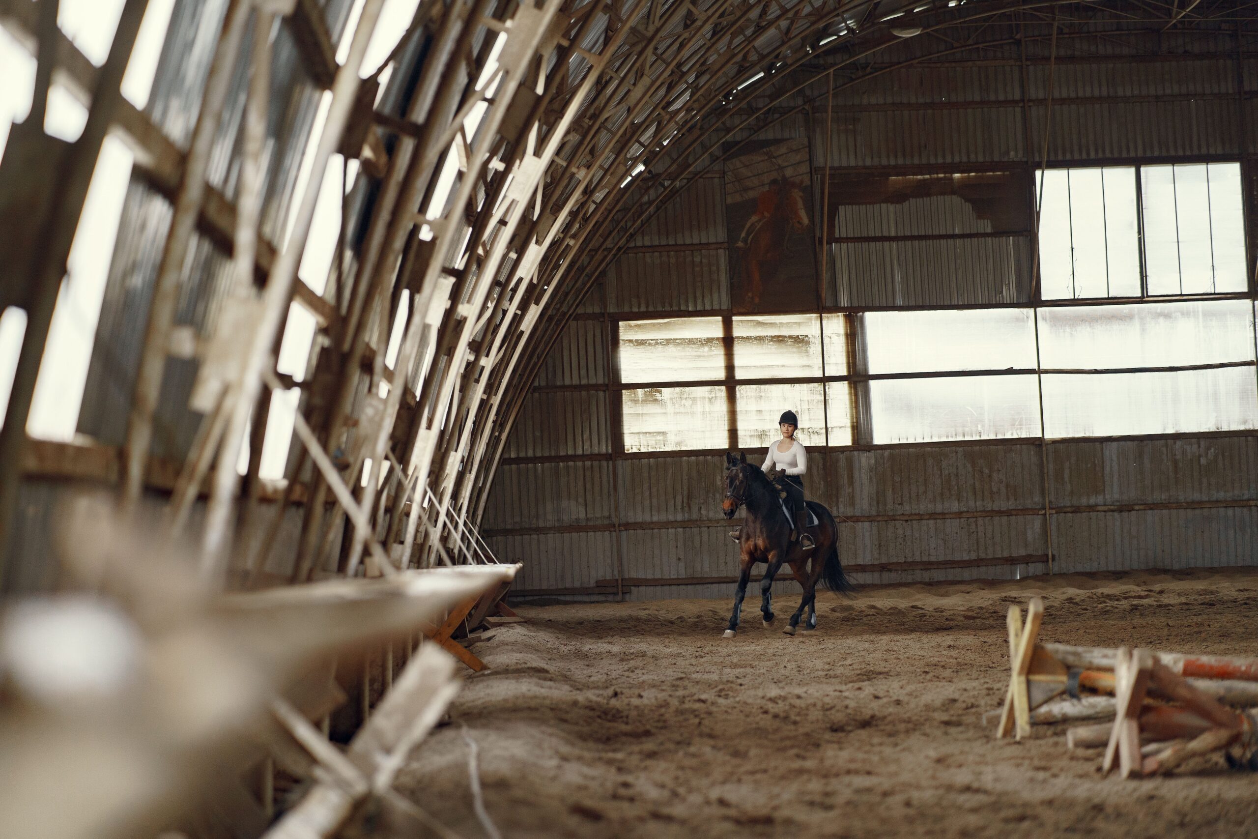 A woman rides a horse in a rustic indoor riding hall surrounded by wooden structures.